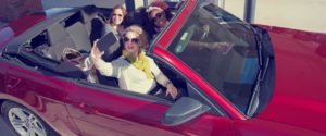 Women driving a convertible and taking a photo.