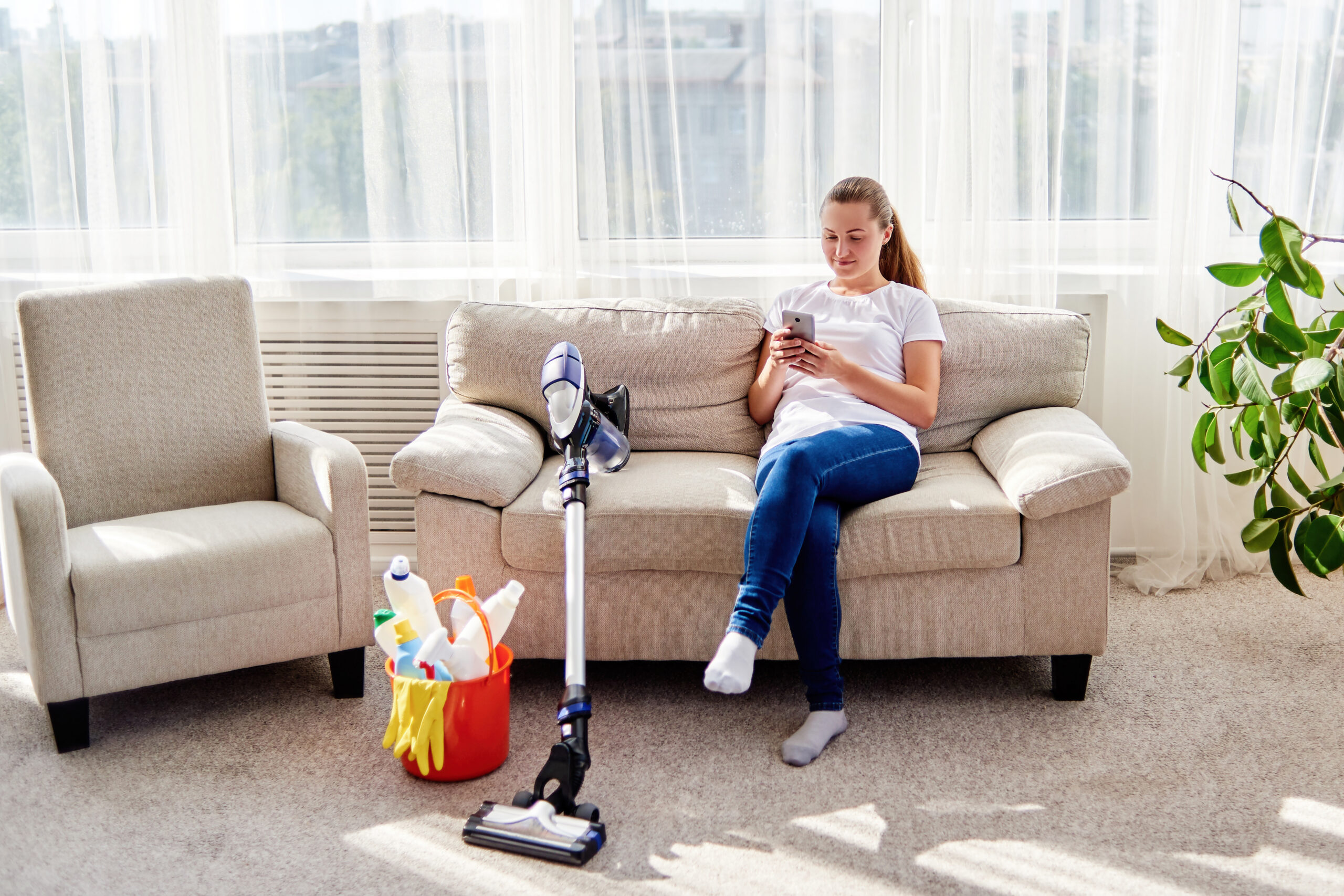 Young woman in white shirt and jeans sitting on sofa and using mobile phone while cleaning at home, copy space. Housework and chores concept. Cleaning appliances